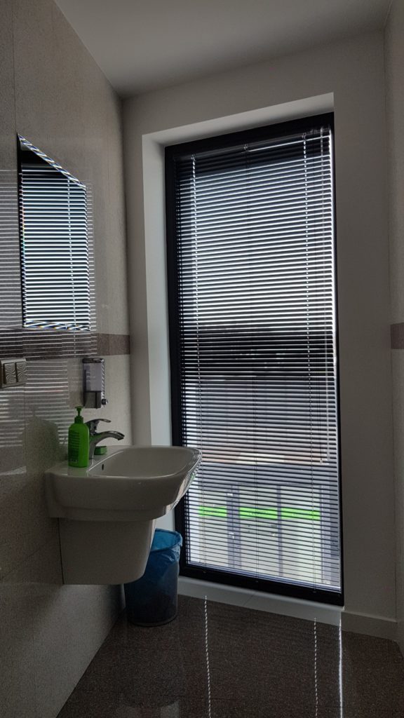 Na zdjęciu widać czarną żaluzję aluminiową zamontowaną na oknie w łazience